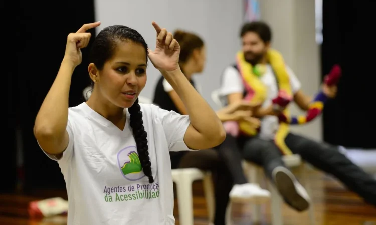 Mulher fazendo interpretação em Libras, com pessoas ao fundo, representando inclusão e acessibilidade.