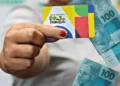 Cartão do Bolsa Família sendo segurado por uma pessoa, com notas de R$ 100 reais visíveis ao fundo. Imagem ilustrativa do programa de assistência social no Brasil.