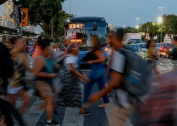 Pessoas apressadas atravessando a rua em meio a um trânsito intenso, ilustrando o cotidiano de trabalho e impostos pesados.