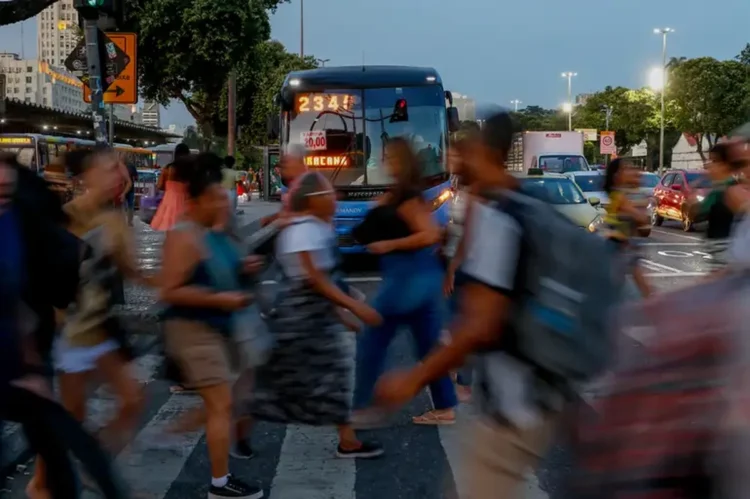 Pessoas apressadas atravessando a rua em meio a um trânsito intenso, ilustrando o cotidiano de trabalho e impostos pesados.