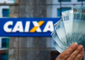 Mão segurando notas de 100 reais em frente à agência da Caixa Econômica Federal.