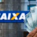 Mão segurando notas de 100 reais em frente à agência da Caixa Econômica Federal.