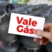 Cartão com os dizeres "Vale Gás" sendo segurado por uma mão, com botijões ao fundo