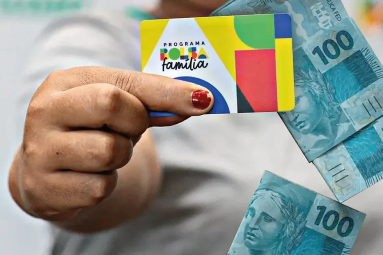 Cartão do Bolsa Família sendo segurado com cédulas de 100 reais ao fundo.