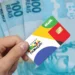 Cartão do Bolsa Família com cédulas de R$ 100 ao fundo.