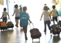 Crianças andando em corredor de escola com mochilas de rodinhas, vista de costas.