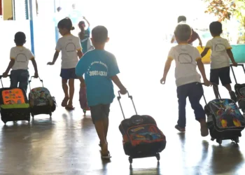Crianças andando em corredor de escola com mochilas de rodinhas, vista de costas.