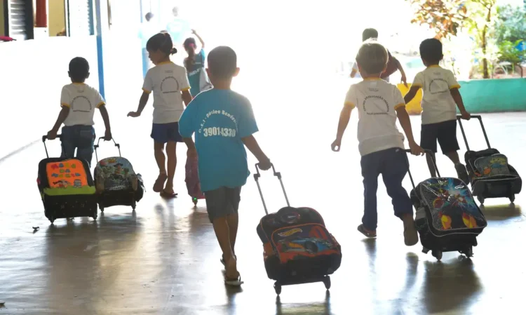 Crianças andando em corredor de escola com mochilas de rodinhas, vista de costas.