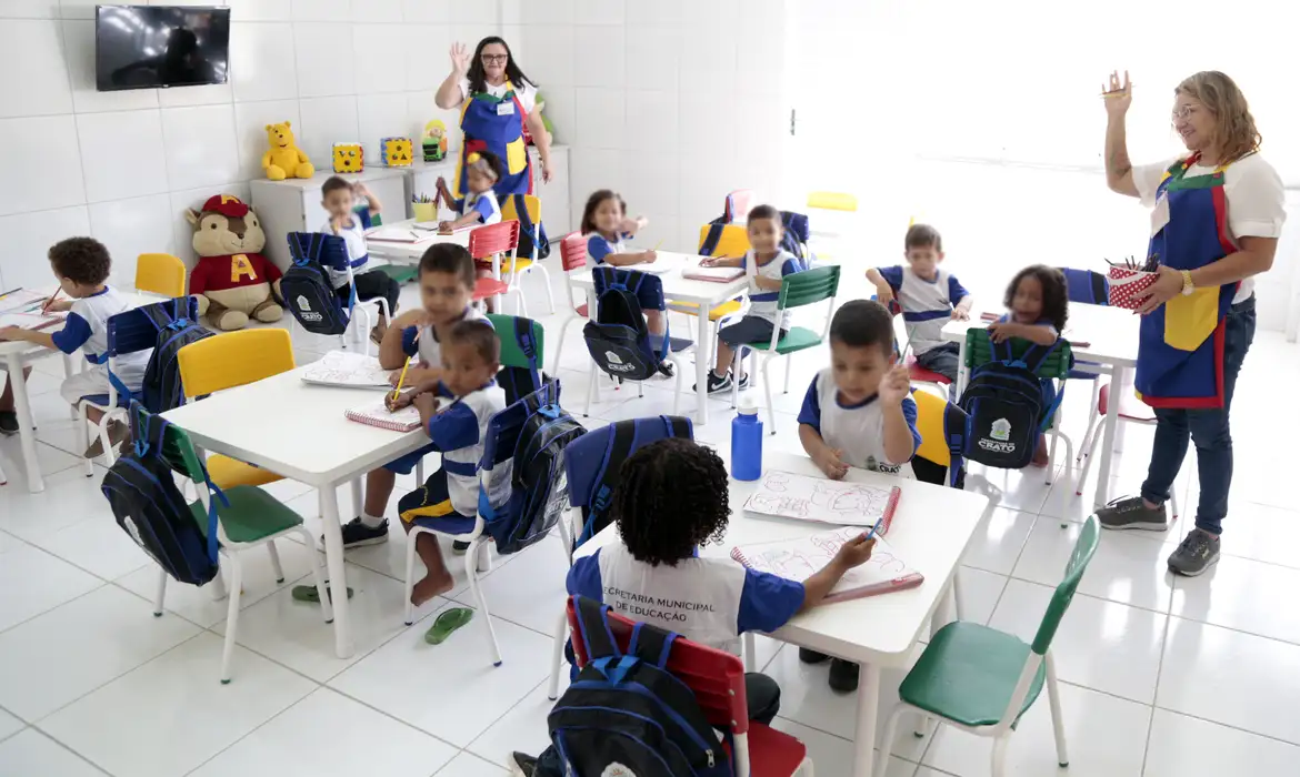 Crianças em sala de aula com professores, sentadas em mesas coloridas, participando de atividade escolar.