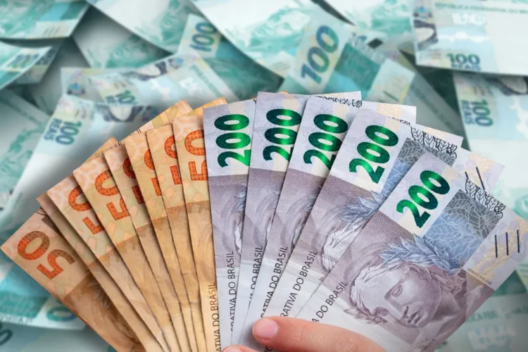 Notas de 50 e 200 reais em destaque com várias cédulas ao fundo.
