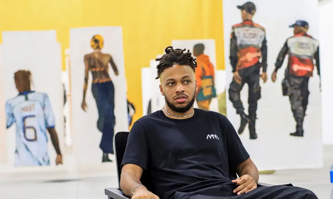 Jovem sentado em cadeira com obras de arte ao fundo.