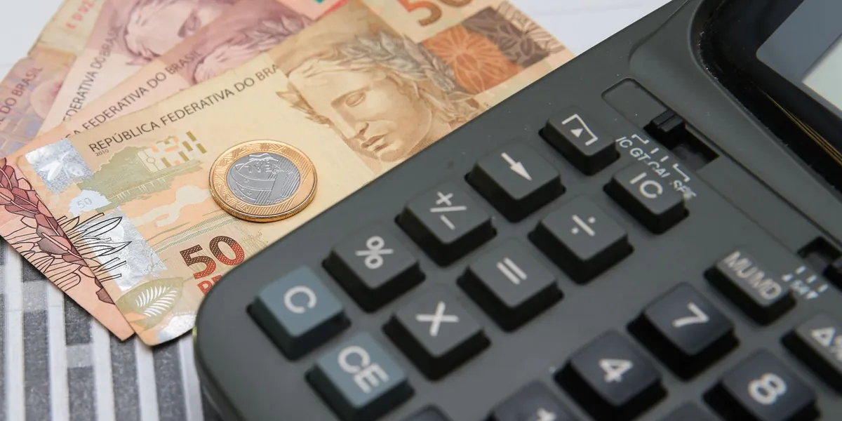 Calculadora ao lado de cédulas e moeda brasileira, simbolizando o peso dos impostos no bolso do trabalhador.