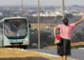 Mulher acenando para ônibus em via urbana com cidade ao fundo