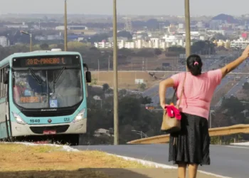 Mulher acenando para ônibus em via urbana com cidade ao fundo