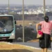 Mulher acenando para ônibus em via urbana com cidade ao fundo