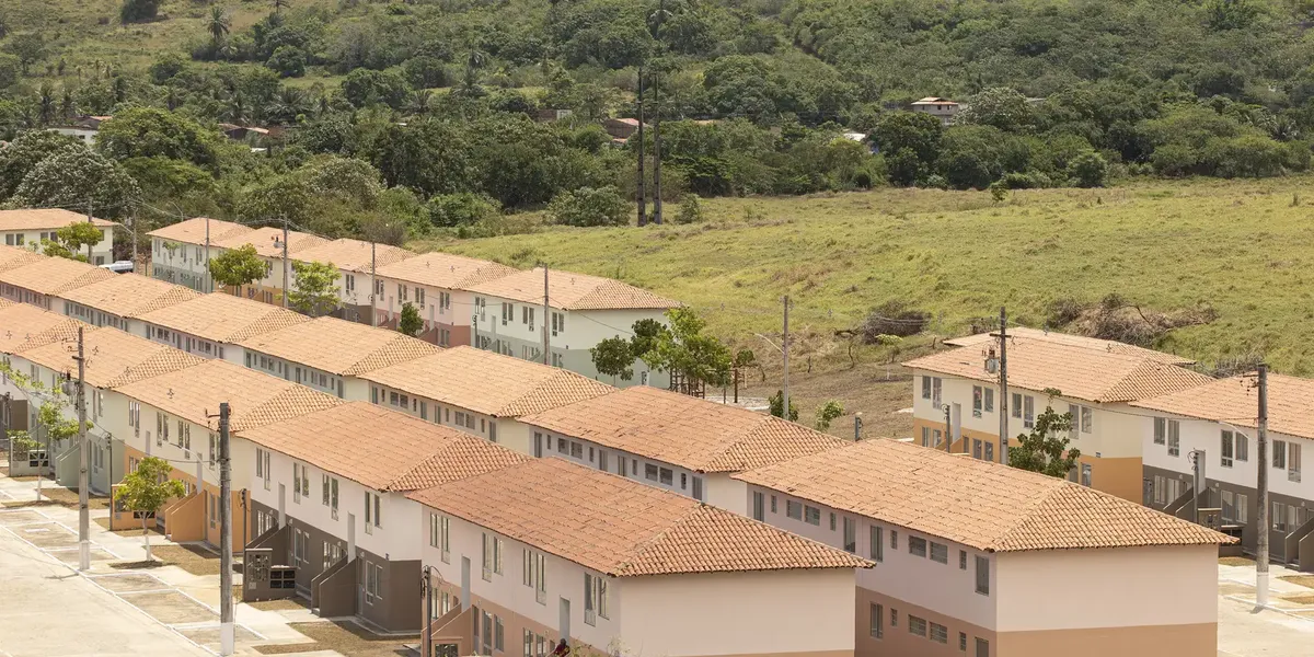 Residencial do programa Minha Casa Minha Vida com casas geminadas em área rural cercada por vegetação.