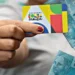 Cartão do programa Bolsa Família sendo segurado por uma mão com esmalte vermelho, ao fundo notas de dinheiro de 100 reais.