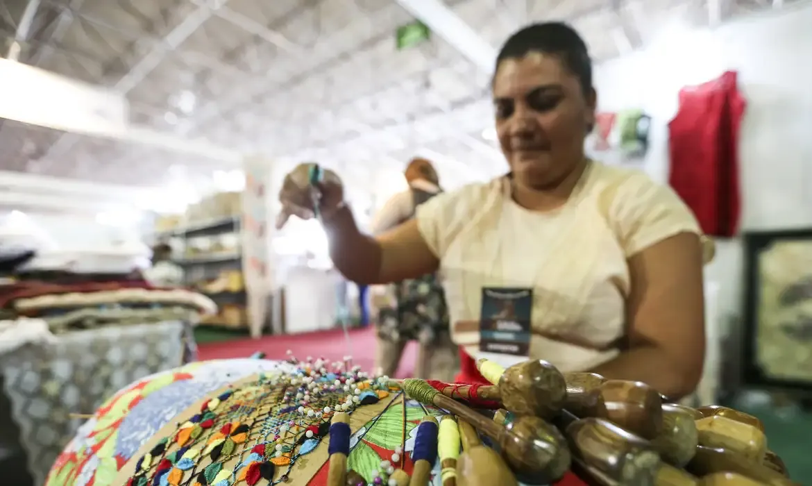 Mulher artesã confeccionando peça artesanal em feira de economia solidária.