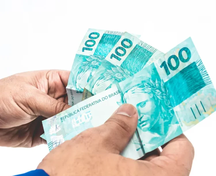 Notas de R$ 100 do Brasil sendo seguradas, ilustrando o valor do 13º salário.