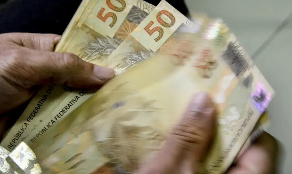 "Imagem de notas de R$ 50 sendo contadas à mão, representando o cálculo dos benefícios.