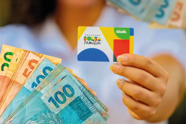 Cartão do programa Bolsa Família sendo segurado, com cédulas de reais visíveis ao fundo