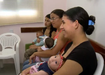 Mães sentadas em sala de espera, cada uma segurando um bebê nos braços.
