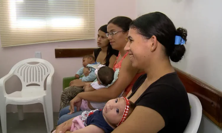 Mães sentadas em sala de espera, cada uma segurando um bebê nos braços.