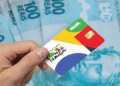 Cartão do Bolsa Família sendo segurado por uma mão com cédulas de 100 reais ao fundo.