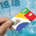 Cartão do Bolsa Família sendo segurado por uma mão com cédulas de 100 reais ao fundo.