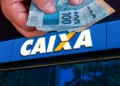 "Pessoa segurando dinheiro na frente de uma agência da Caixa Econômica Federal"