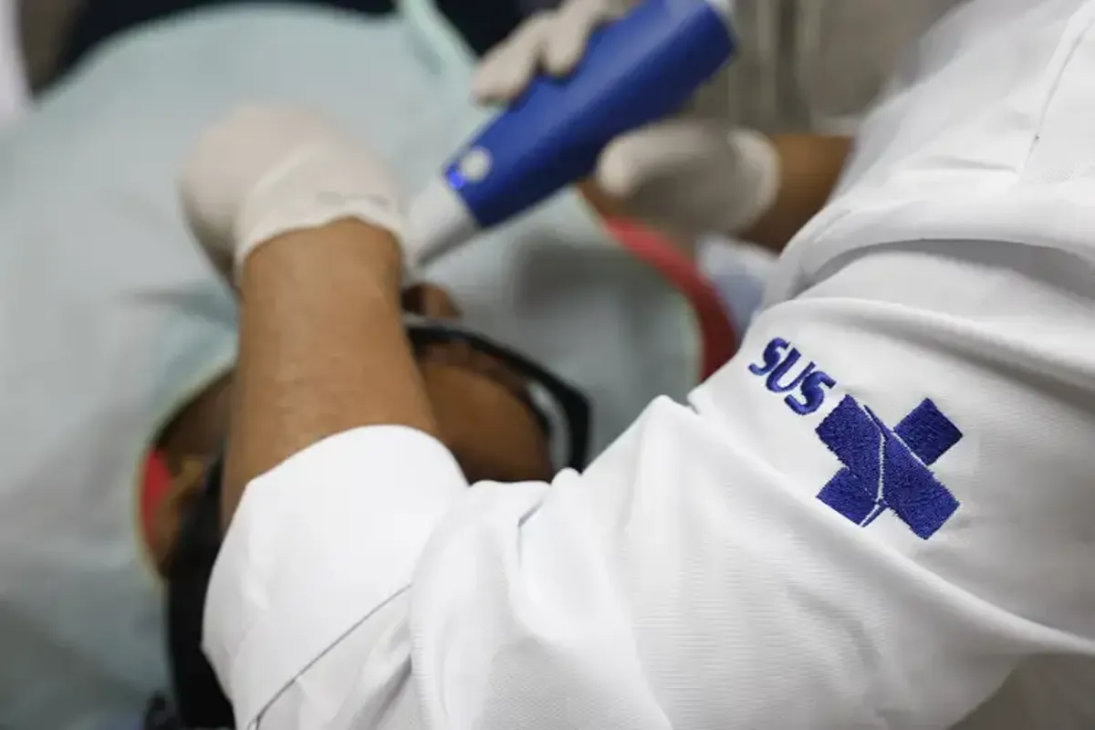 Imagem de um profissional de saúde atendendo um paciente no SUS, com o logo do SUS visível na camisa.