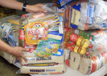 Imagem de cestas básicas sendo preparadas para distribuição, com produtos como arroz, óleo e macarrão.