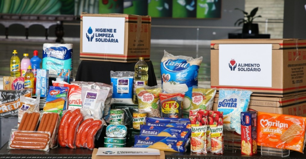 Imagem de cestas básicas contendo alimentos e produtos de higiene, destacando as caixas 'Alimento Solidário' e 'Higiene e Limpeza Solidária', com diversos itens como arroz, óleo, salsichas, macarrão, produtos de limpeza e higiene.