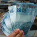 Mão segurando notas de 100 reais sobre pilhas de dinheiro, representando crédito consignado.