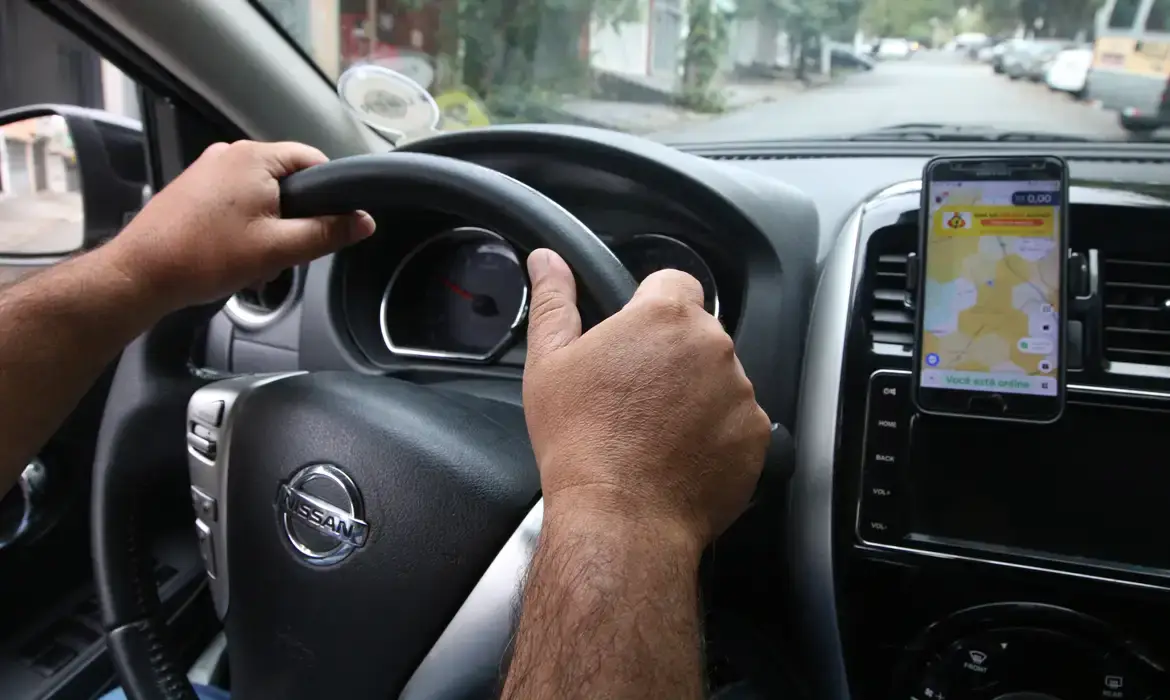 Mãos de motorista de aplicativo no volante com aplicativo de transporte aberto no celular