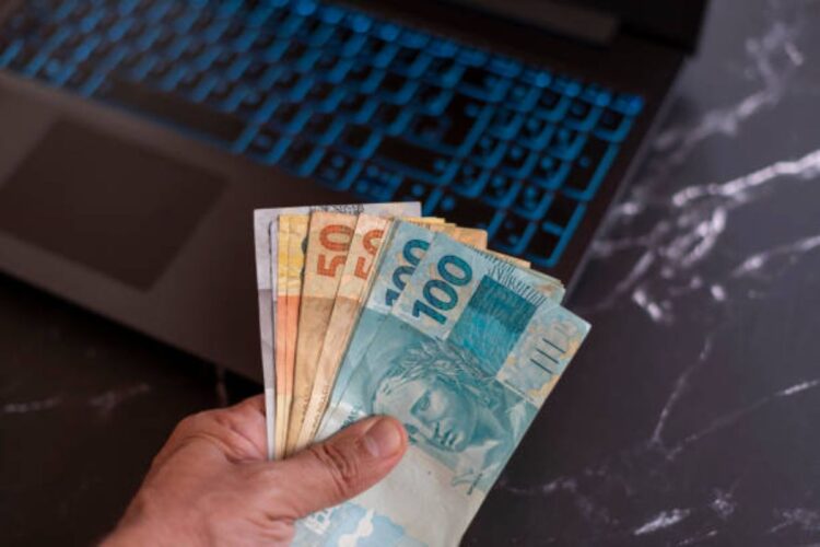 Mão segurando notas de real em frente a um notebook