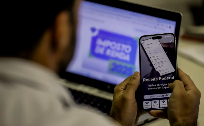Pessoa acessando o aplicativo da Receita Federal no celular enquanto visualiza informações sobre o Imposto de Renda no computador.