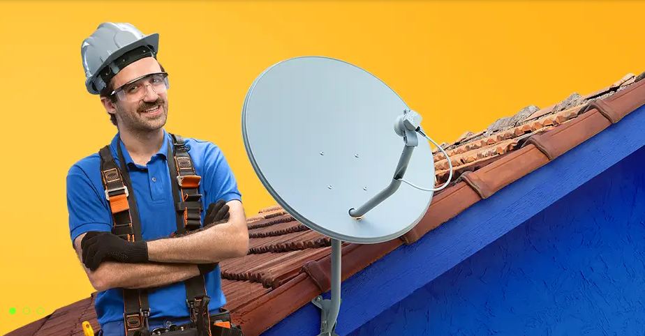 Homem sorridente com capacete e equipamentos de segurança, posando ao lado de uma parabólica digital instalada no telhado de uma casa colorida.
