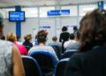 Sala de espera com atendentes e pacientes no INSS, com tela indicando senhas de atendimento
