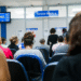 Sala de espera com atendentes e pacientes no INSS, com tela indicando senhas de atendimento