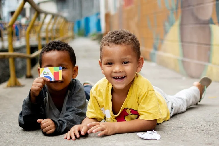 Crianças sorrindo deitadas na calçada, uma delas segurando cartão do Bolsa Família