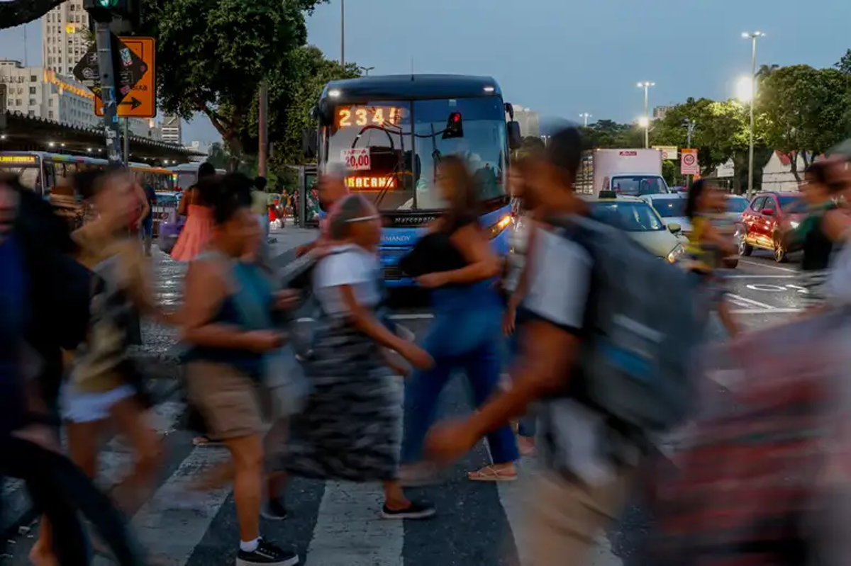 Pedestres cruzando a rua com um ônibus em movimento, destacando o pagamento da 1ª parcela do 13º salário em julho.