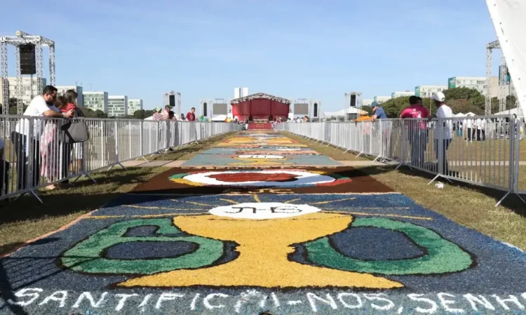Tapete colorido de Corpus Christi em um evento ao ar livre, com desenhos religiosos e pessoas ao fundo.