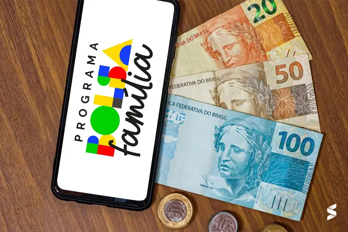 Imagem de um smartphone com o logo do Programa Bolsa Família e cédulas de dinheiro ao fundo, representando o auxílio financeiro.