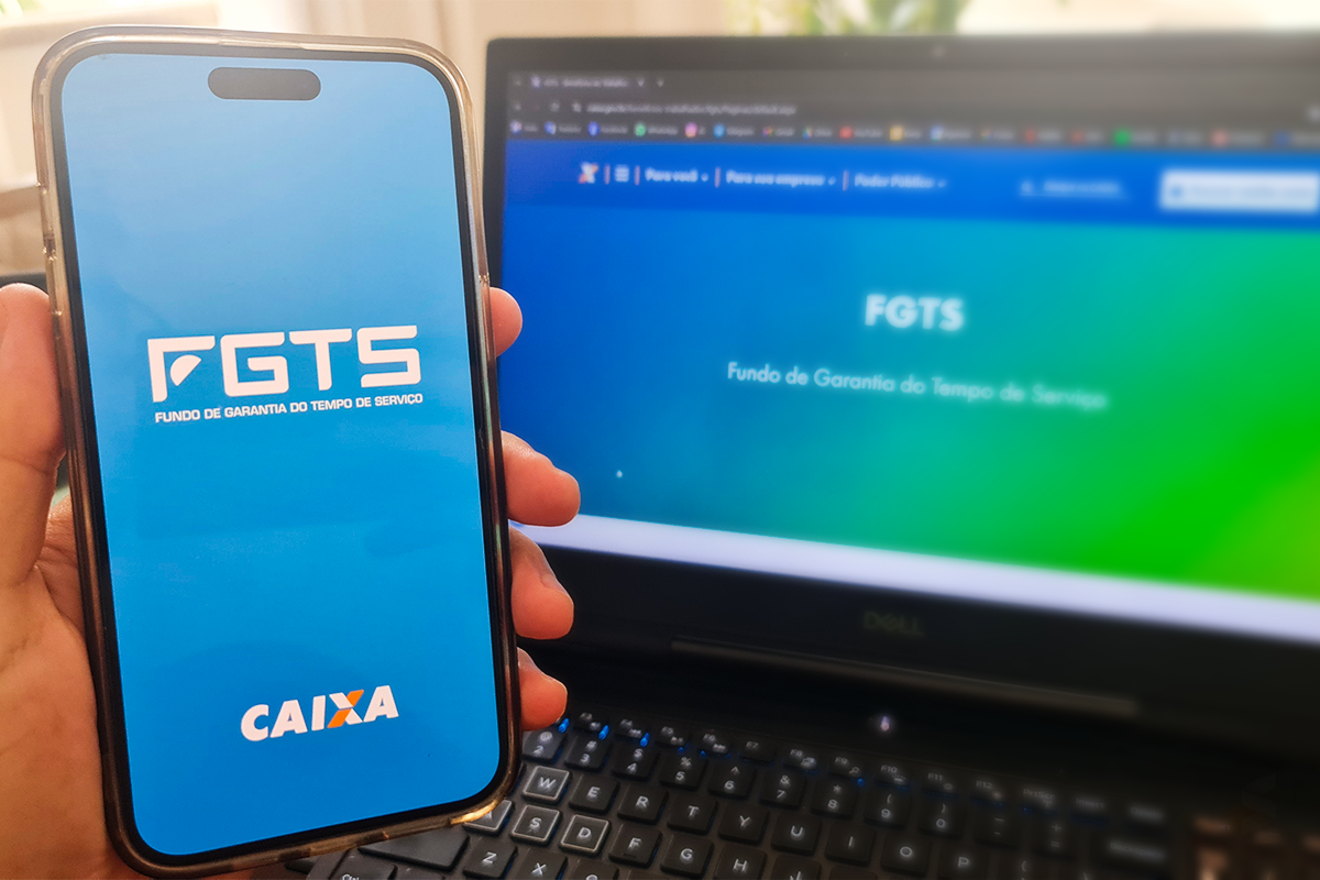 Pessoa segurando um celular com o logo do FGTS, enquanto um laptop exibe a página do FGTS, simbolizando os canais de atendimento para quitar dívidas com o FGTS.