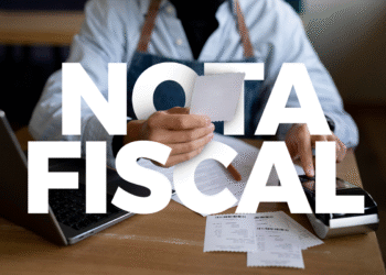 Pessoa segurando nota fiscal com computador e comprovantes sobre a mesa