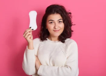 Mulher jovem sorrindo e segurando um absorvente higiênico em frente a um fundo rosa.