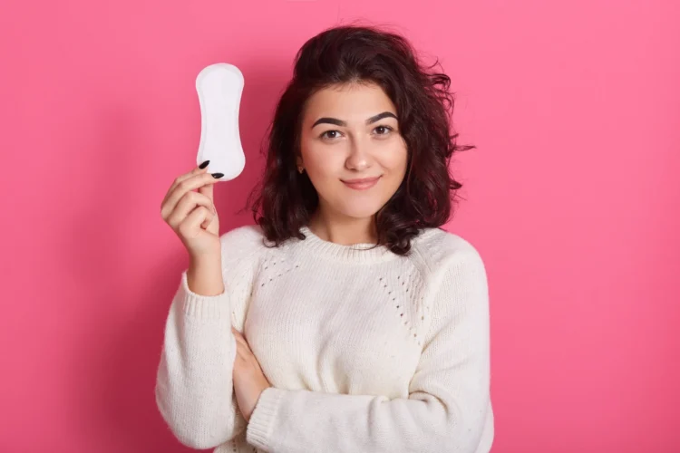 Mulher jovem sorrindo e segurando um absorvente higiênico em frente a um fundo rosa.