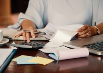 Pessoa organizando contas com calculadora, laptop e boletos sobre a mesa