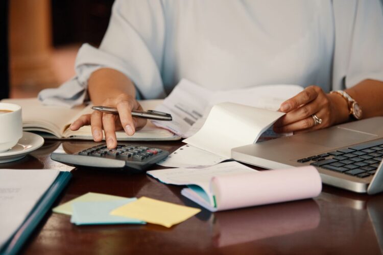 Pessoa organizando contas com calculadora, laptop e boletos sobre a mesa